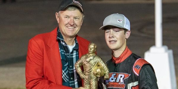 A young race car driver receives a golden trophy from a man in a red jacket.