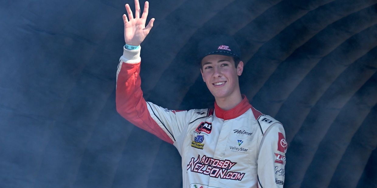 Race car driver waving and smiling in his racing suit and cap.