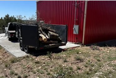 Trailer loaded with cut logs parked beside a red building on a sunny day.