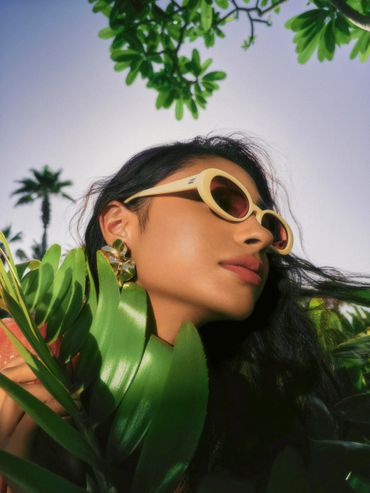 Woman wearing yellow sunglasses surrounded by green leaves under clear sky.