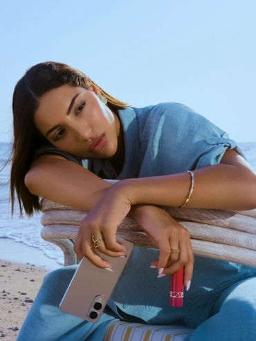 Woman in blue shirt leaning on a chair at the beach, holding a phone and lipstick.