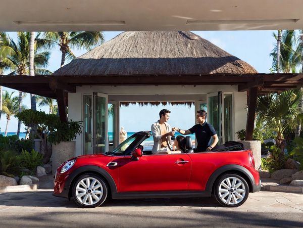Two men exchanging car keys beside a red convertible under a tropical hut.