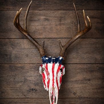 Deer skull with American flag painted on it, mounted on wooden wall.