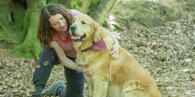 A woman kneeling and hugging a happy golden retriever in a forest.