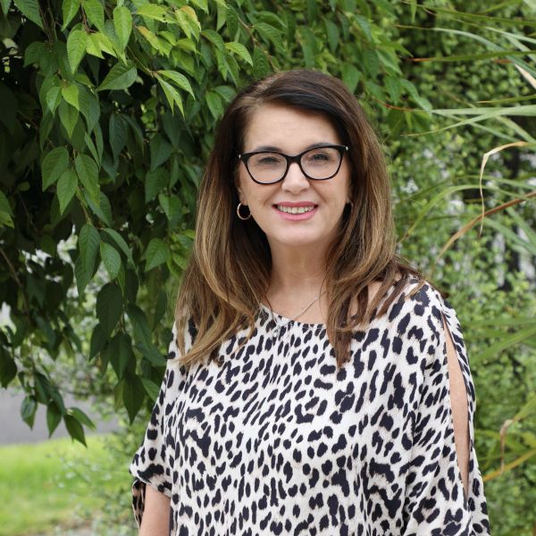 Woman with glasses smiling outdoors in a spotted blouse.