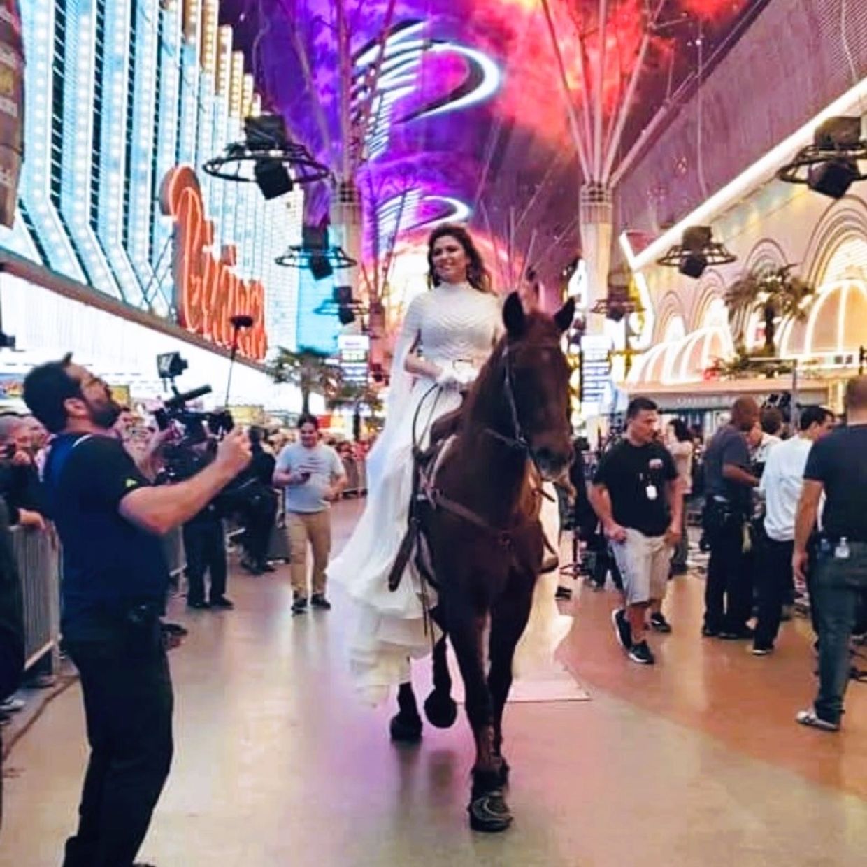 A woman in a white dress rides a horse through a vibrant, crowded street at night.