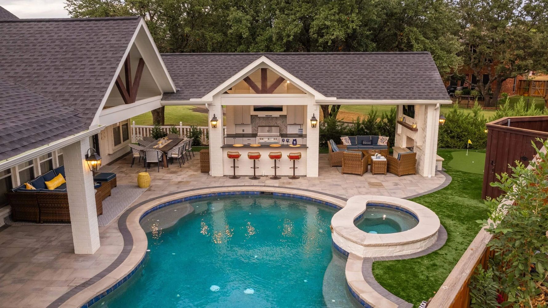 Luxurious backyard with pool, hot tub, outdoor kitchen, and seating areas under a covered patio.