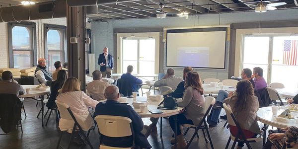 A group attending a presentation in a spacious room with large windows and an American flag.