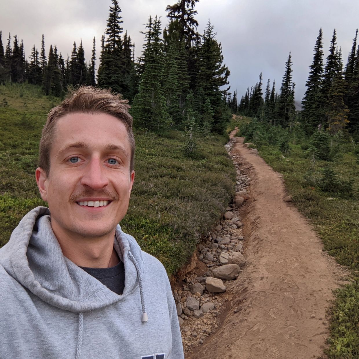 A man in a gray hoodie smiles on a forest hiking trail under a cloudy sky.