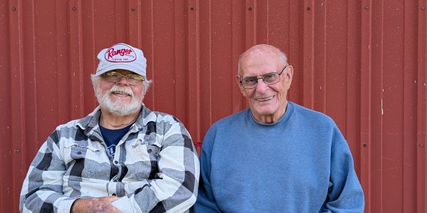 Two elderly men smiling in front of a red wall.