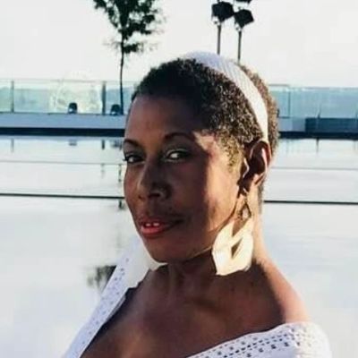 Woman posing outdoors by water, wearing large earrings and a white top.