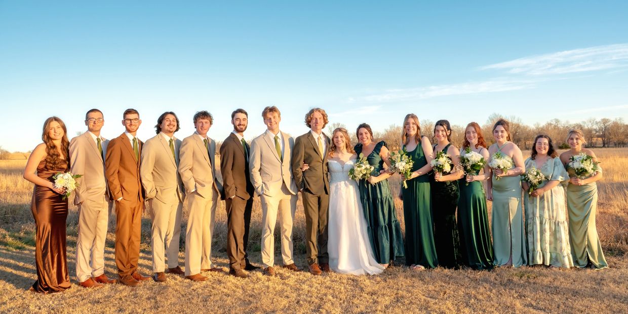 A large wedding party poses outdoors in formal attire under a clear sky.