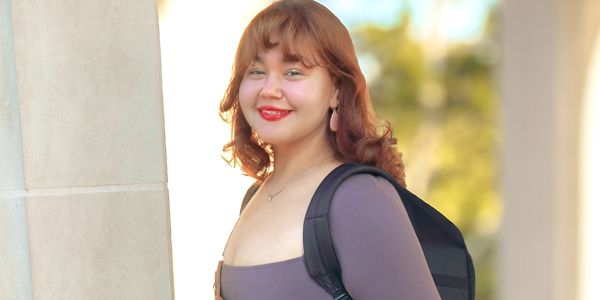 Young woman with a camera poses outdoors by a stone pillar.