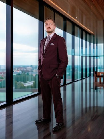 Man in a burgundy suit standing by a large window.