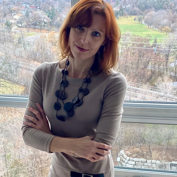 Woman with red hair standing by the window with arms crossed.