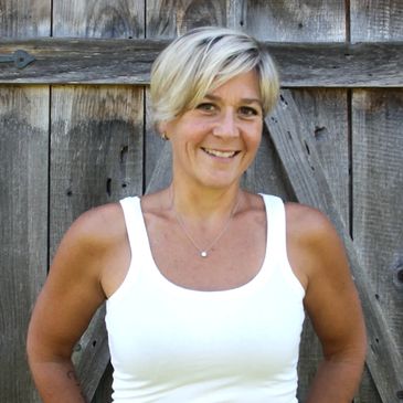 Smiling woman in a white tank top stands in front of a wooden fence.