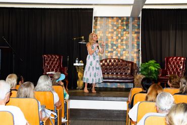 Woman speaking on stage to an audience seated in chairs.