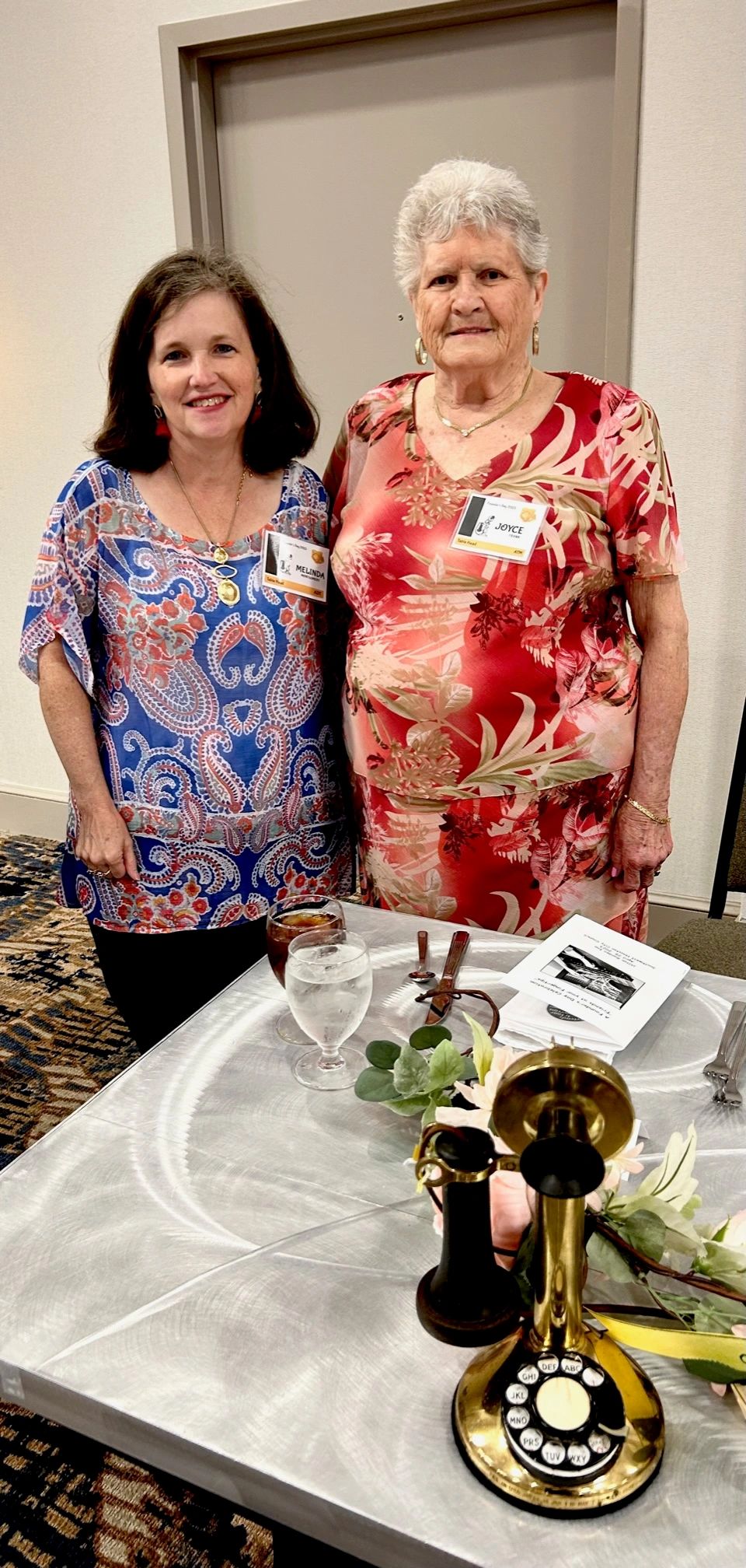 Two women smiling at a social event with an antique telephone on the table.