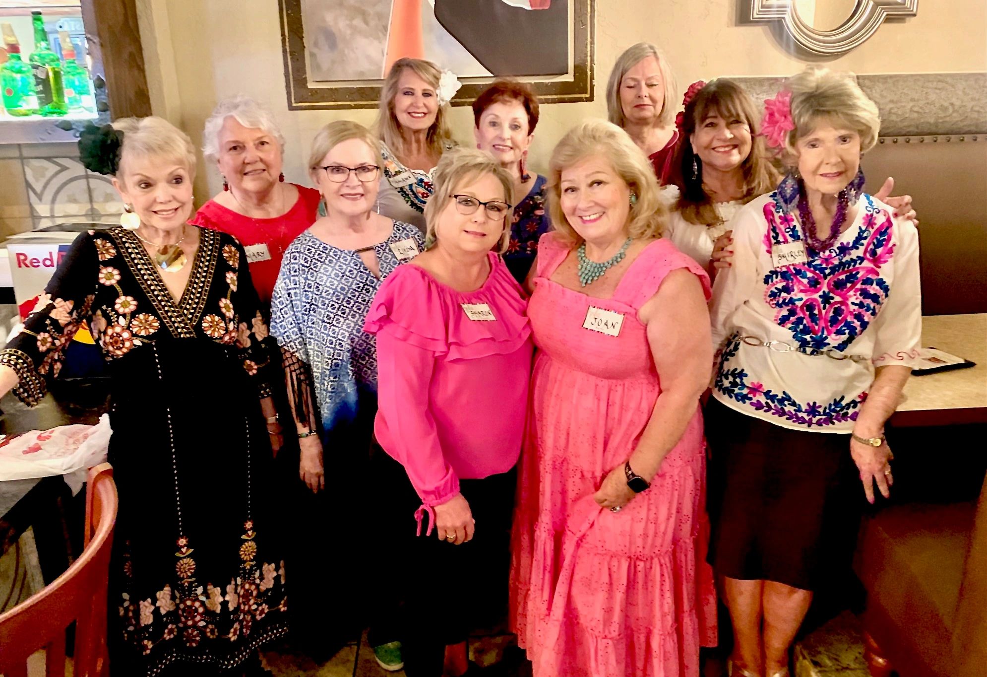 Group of smiling older women posing together indoors with name tags and floral accessories.