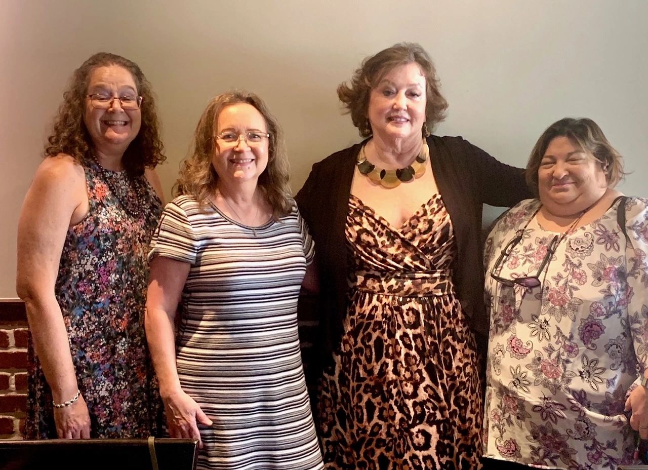 Four smiling women posing together indoors in casual and dressy outfits.