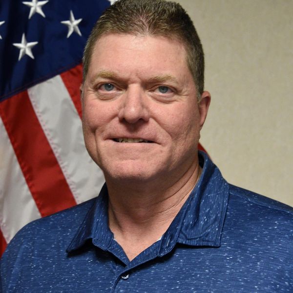 Man in blue shirt standing in front of an American flag.