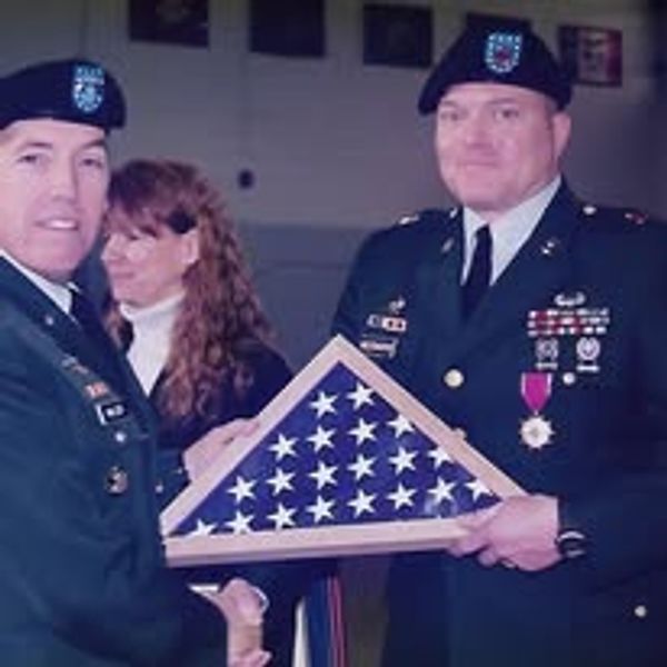 Two military personnel exchanging a folded American flag during a ceremony.