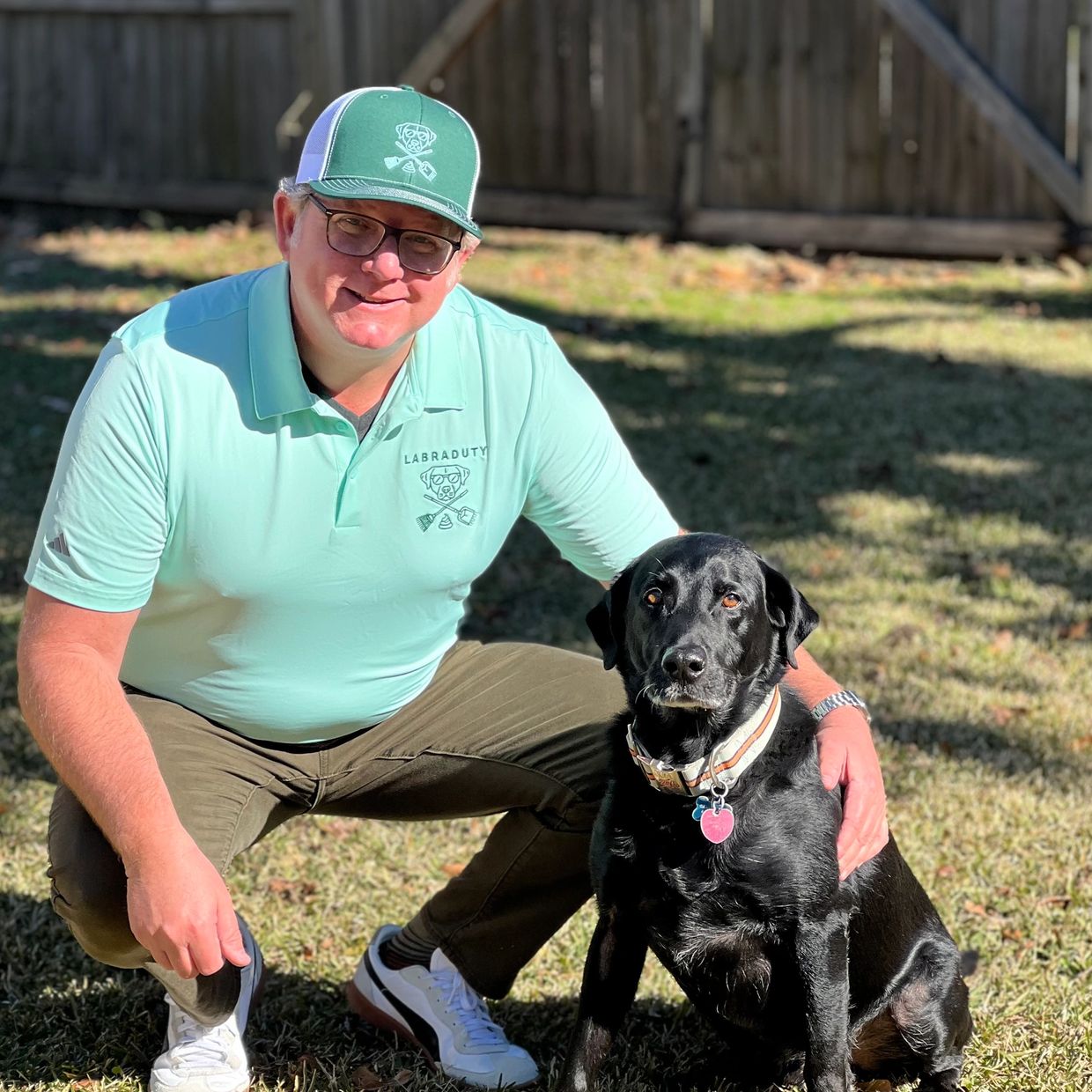 Man in green shirt and cap kneels beside black dog outdoors.