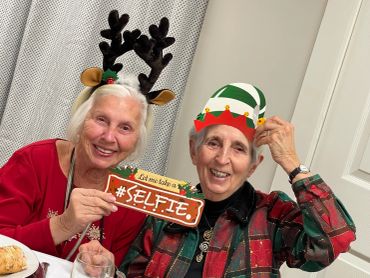 Two elderly women smiling with festive holiday headbands and a selfie sign.