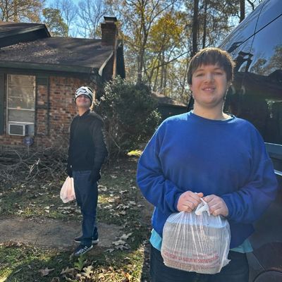 Two men are standing in front of a home. They are holding plastic bags with packed meals in them.