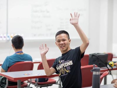 An excited young man with his hands in the air. He is seated in a school setting. He is smiling.