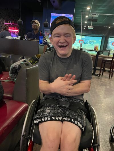 A man in a wheelchair is brightly smiling and wearing a backwards baseball cap.