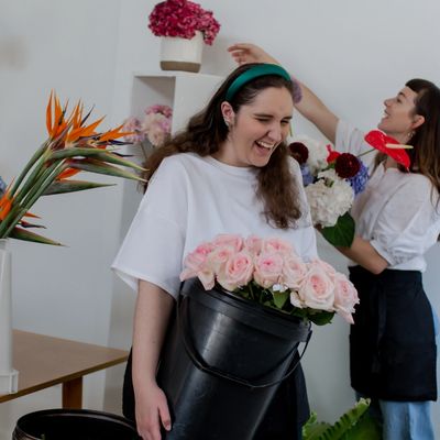 Two women are working in a flower shop. One woman is holding a bucket of pink roses. 