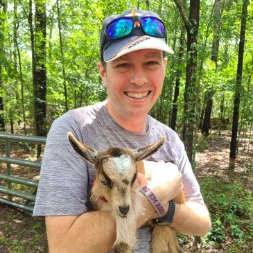 A man is closely holding a baby goat. He is wearing a baseball cap.
