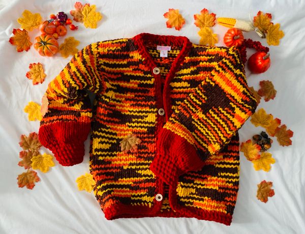 Hand-knit autumn-themed sweater surrounded by fall foliage and mini pumpkins.