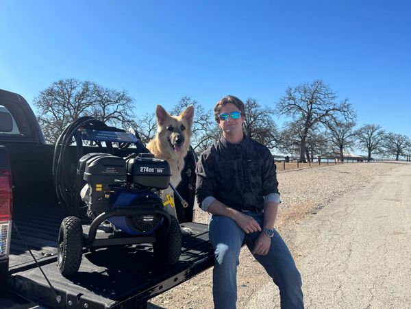 Man with sunglasses and German shepherd sitting on a truck bed next to a pressure washer.