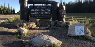 Entrance sign to Grand Canyon National Park surrounded by stones and trees.