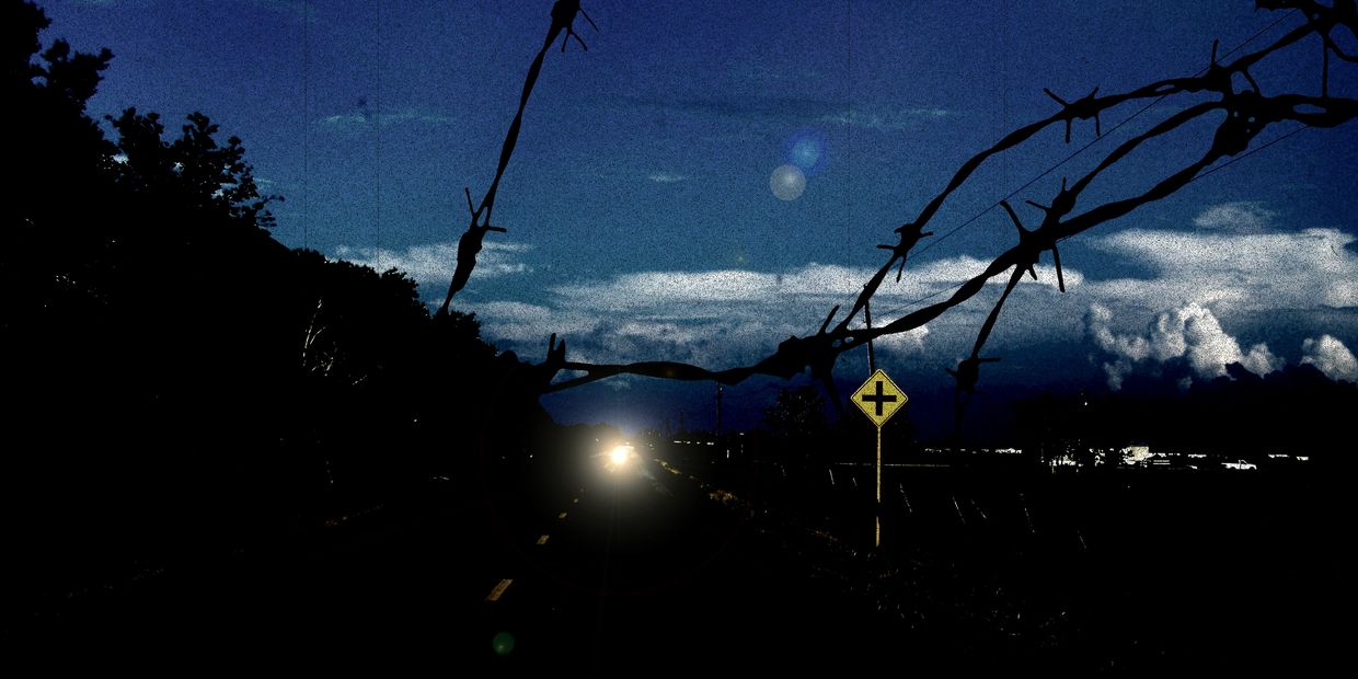 Headlights shine through barbed wire on a dark road at dusk.