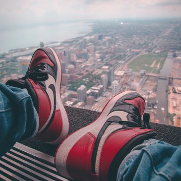 Person wearing black and red Jordan shoes while sitting in a high rise building, overlooking a city.