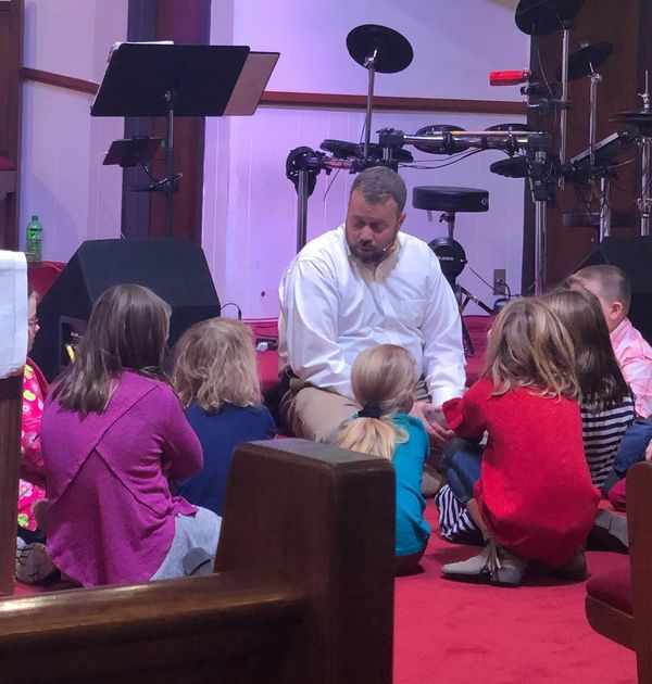 Pastor seated and talking to a group of young children seated in semi-circle on the floor of the san