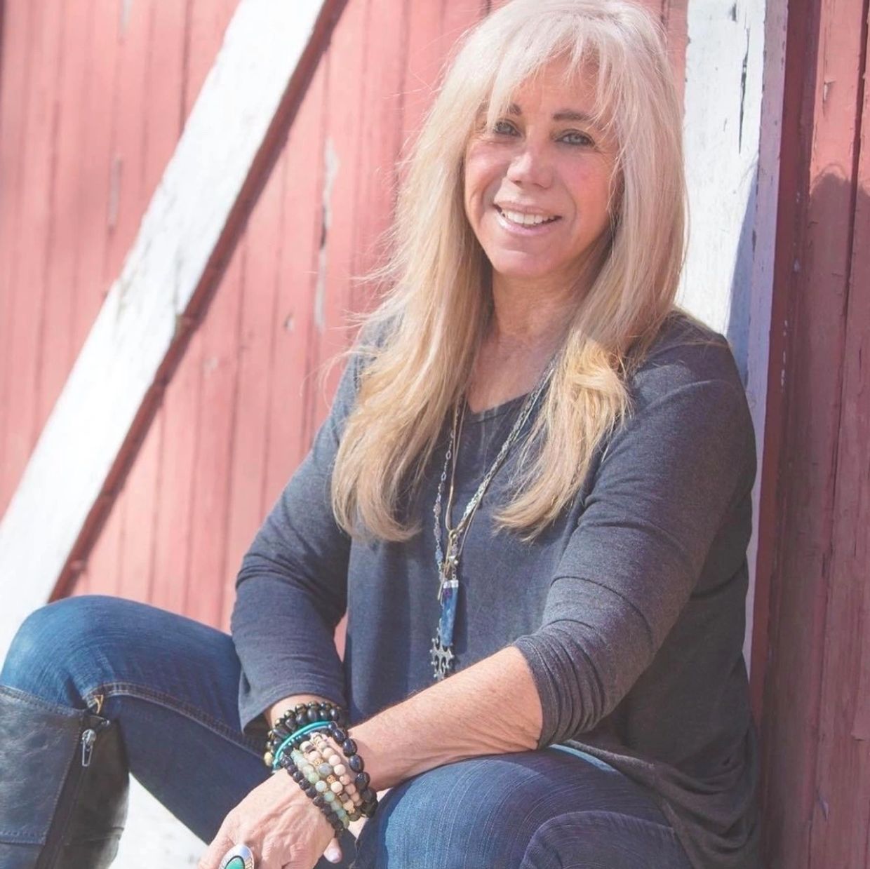 Smiling woman with blonde hair sitting outdoors by a rustic red wall.