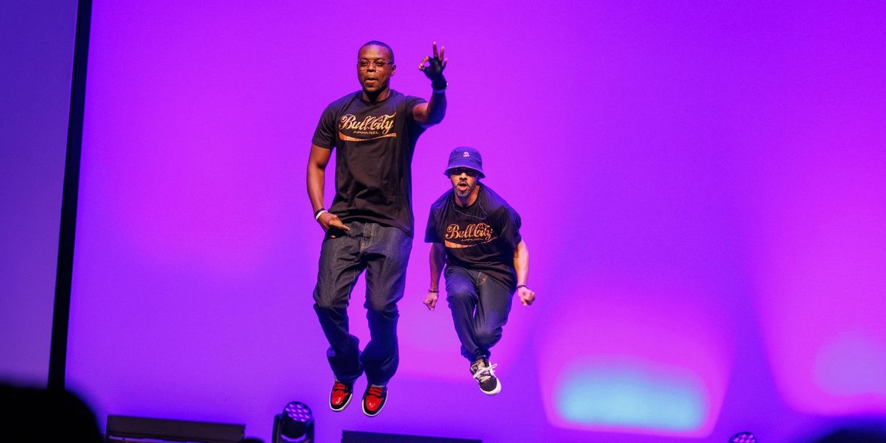 Two men jump energetically on stage against a vibrant purple backdrop.