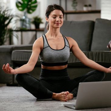 A yoga student in her living room practicing her meditation pose in front of her computer.