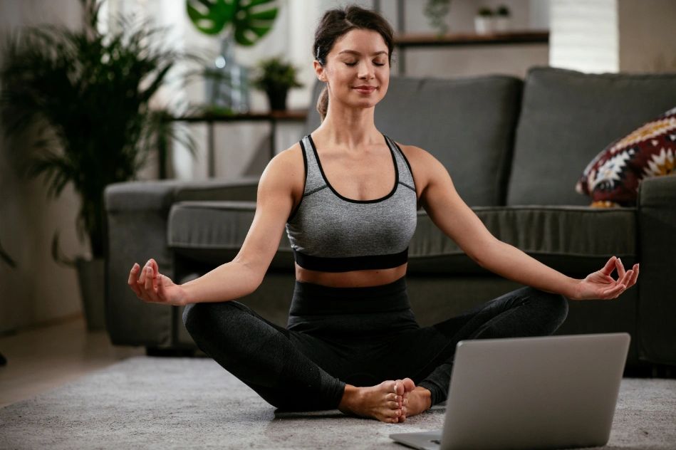 A yoga student sat on the floor of her living room in from of a computer in her meditation pose.