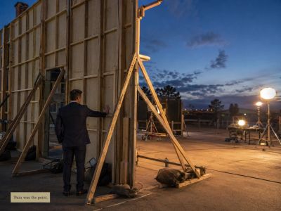 A business-dressed man stands behind a movie-set facade at dusk, quietly confronting the wooden brac
