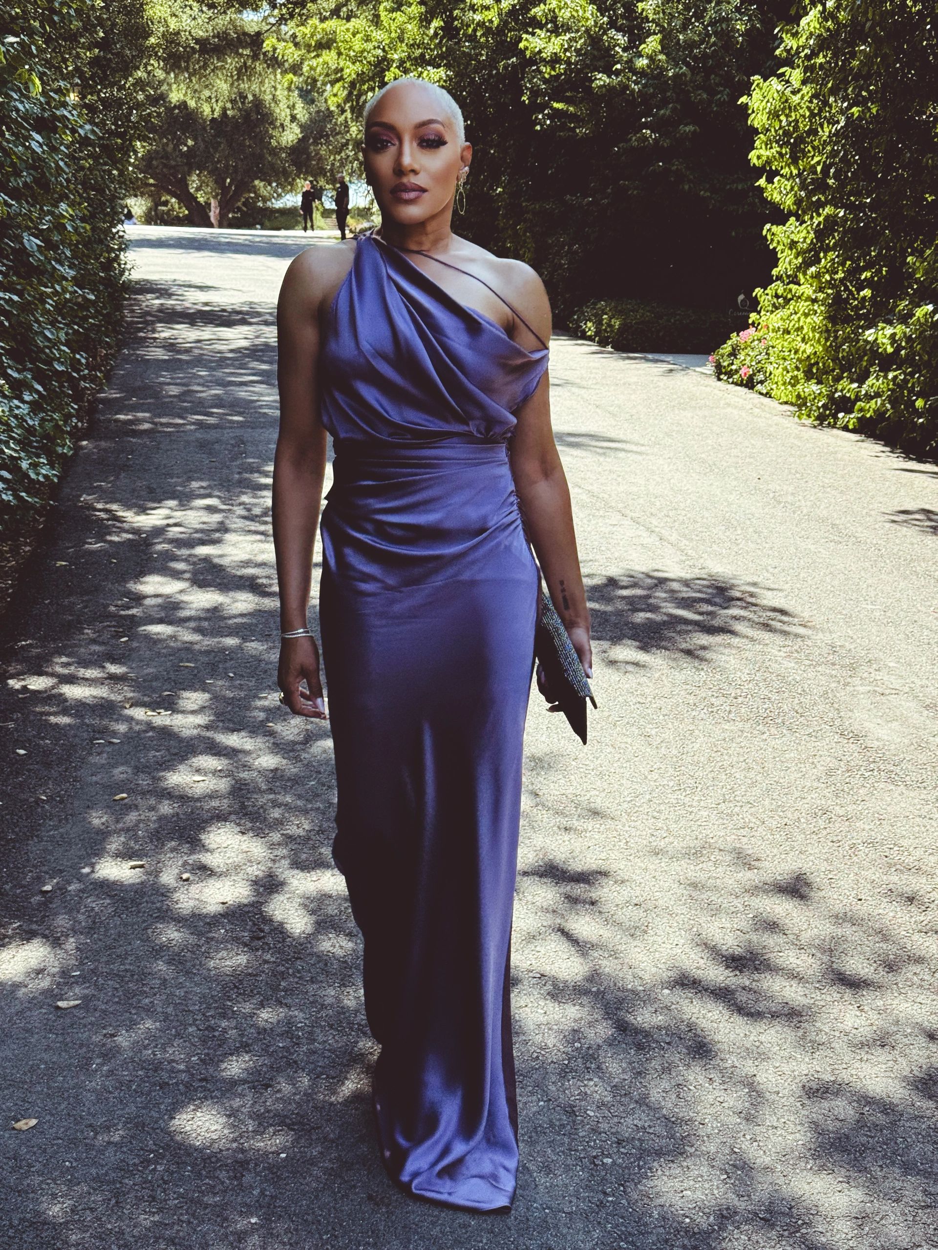Elegant woman in a sleek purple gown walking on a sunlit path.