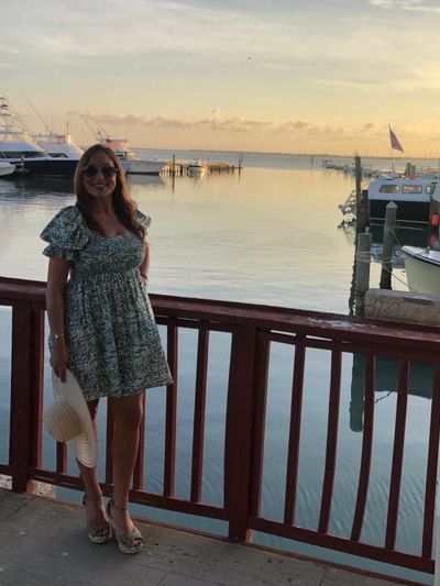 Woman in floral dress standing by marina railing at sunset, holding a hat.