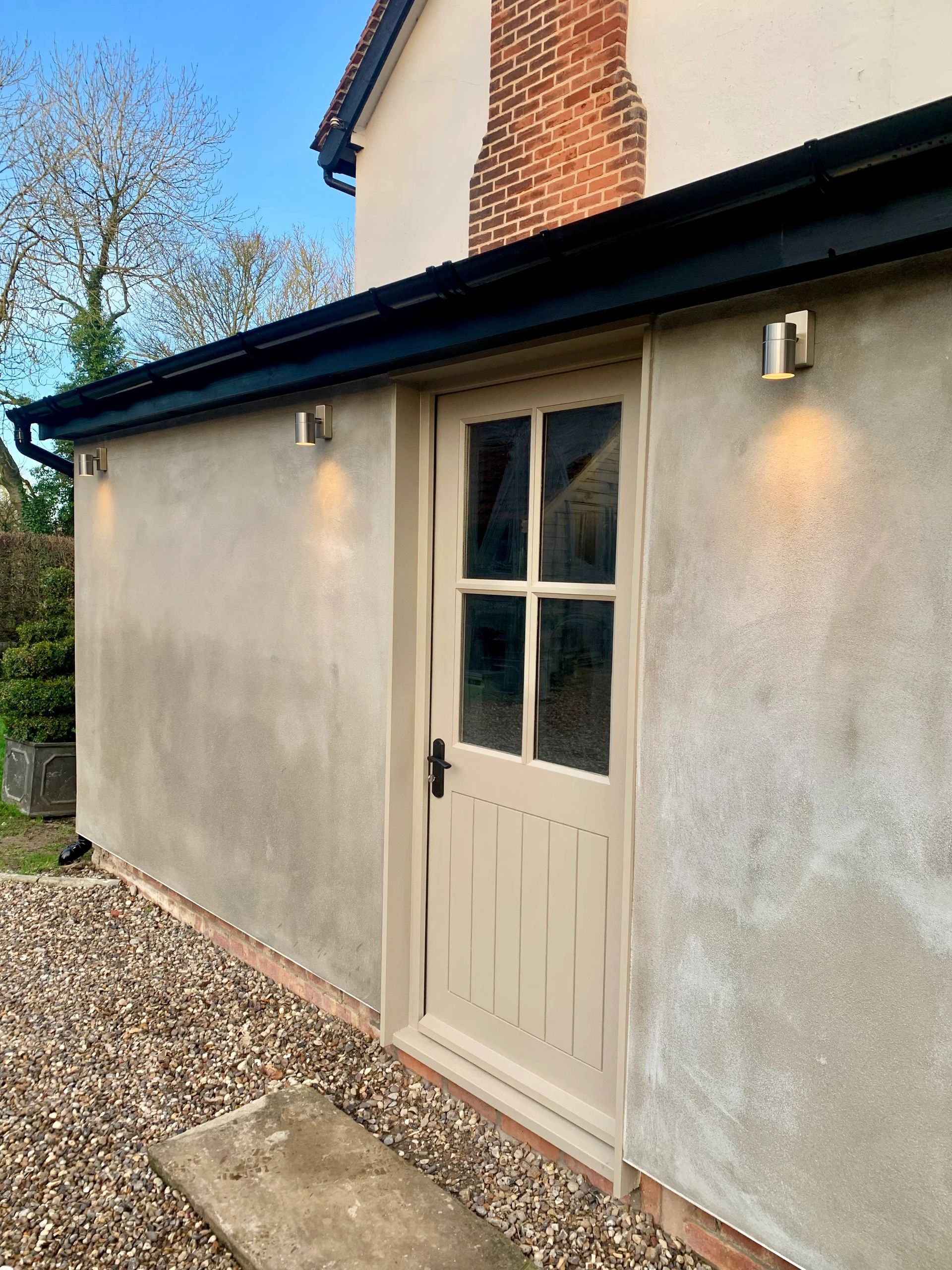 Exterior view of a beige door with glass panels and wall-mounted lights on a grey wall.