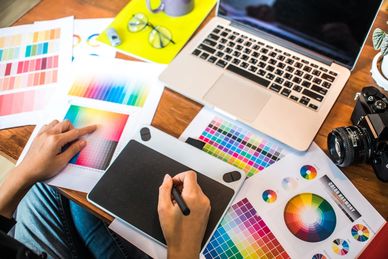 Designer working with color palettes and a graphics tablet beside a laptop and camera.