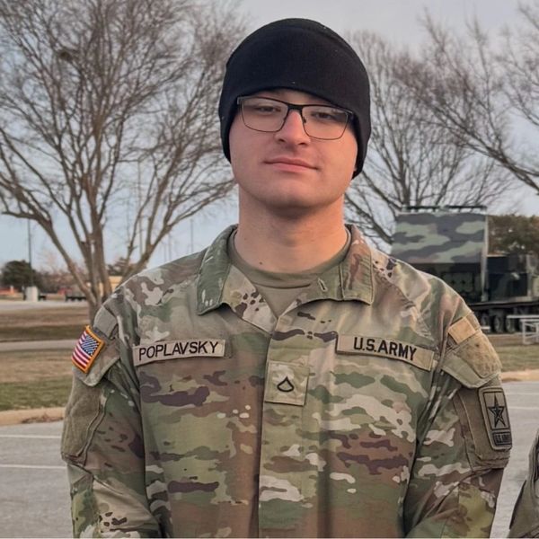 U.S. Army soldier in camouflage uniform with glasses and black beanie outdoors.