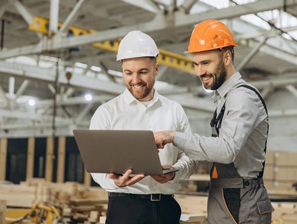 Two men in hard hats review a laptop in a warehouse, collaborating on a project.


